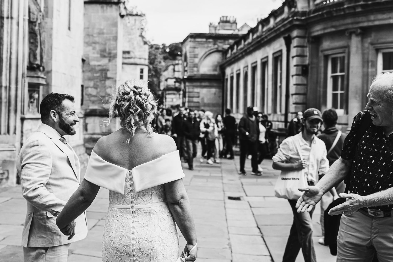Black and white wedding photo of bride and groom outside historic building with guests nearby.