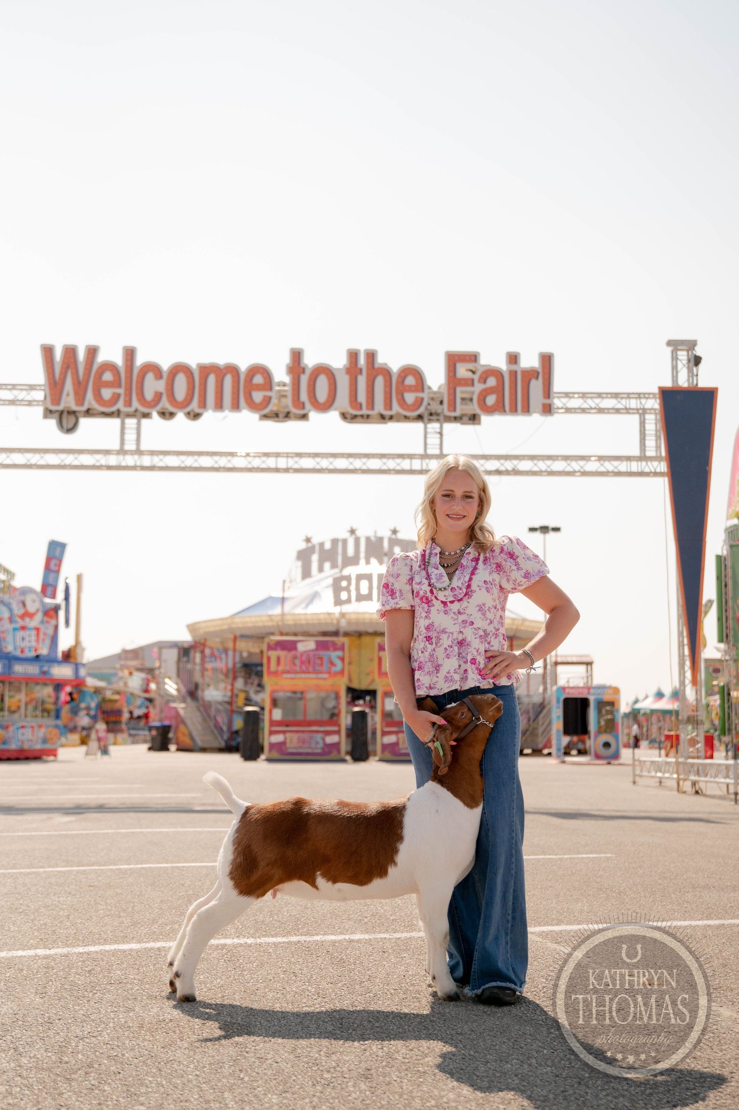 Woman posing with a goat under a Welcome to the Fair sign at a sunny county fair.