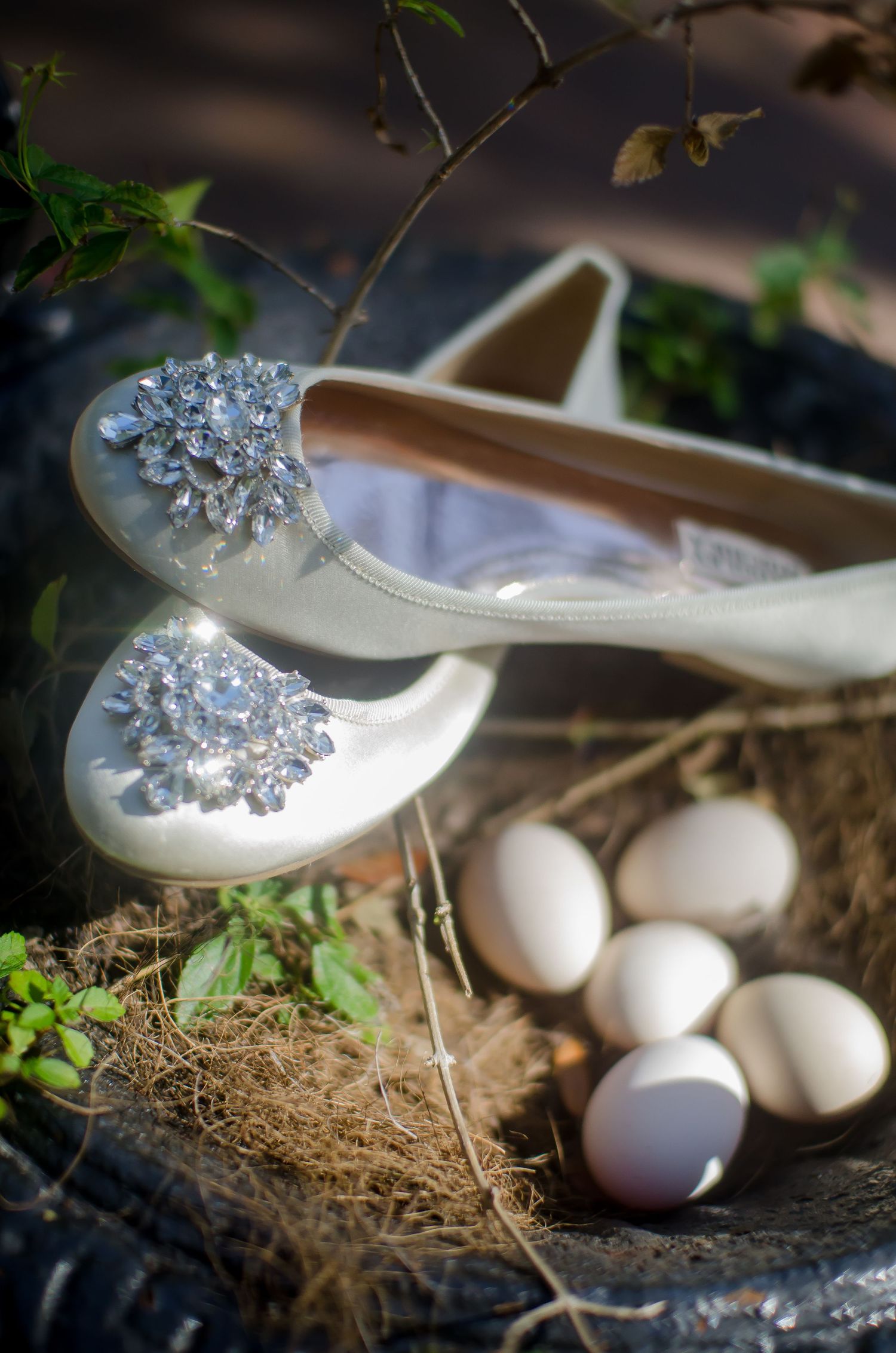 White bridal ballet flats with crystal brooch embellishments displayed beside a bird nest with white eggs outdoors.