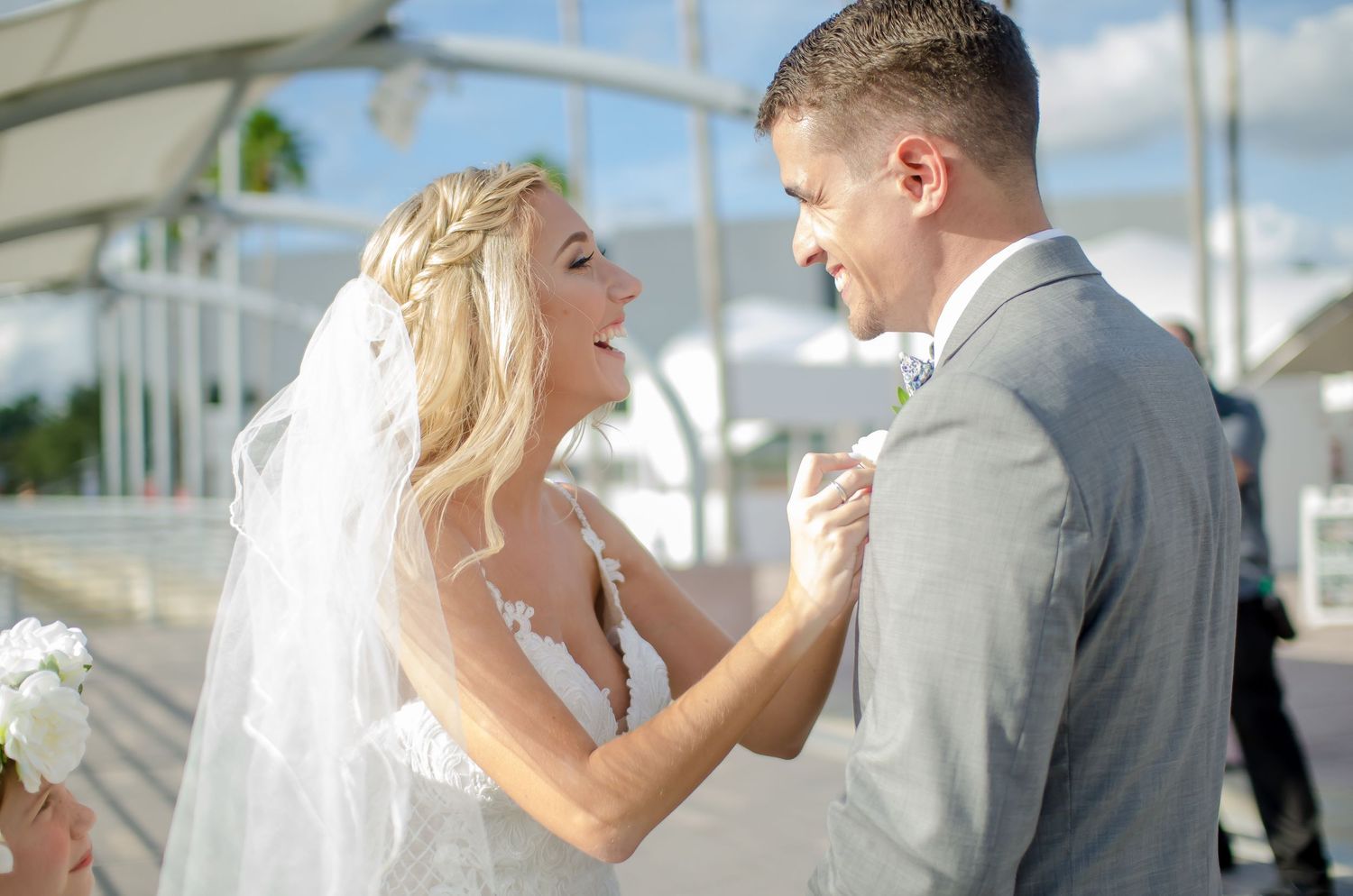 Bride in lace wedding gown and veil smiles at groom in gray suit during outdoor wedding ceremony.