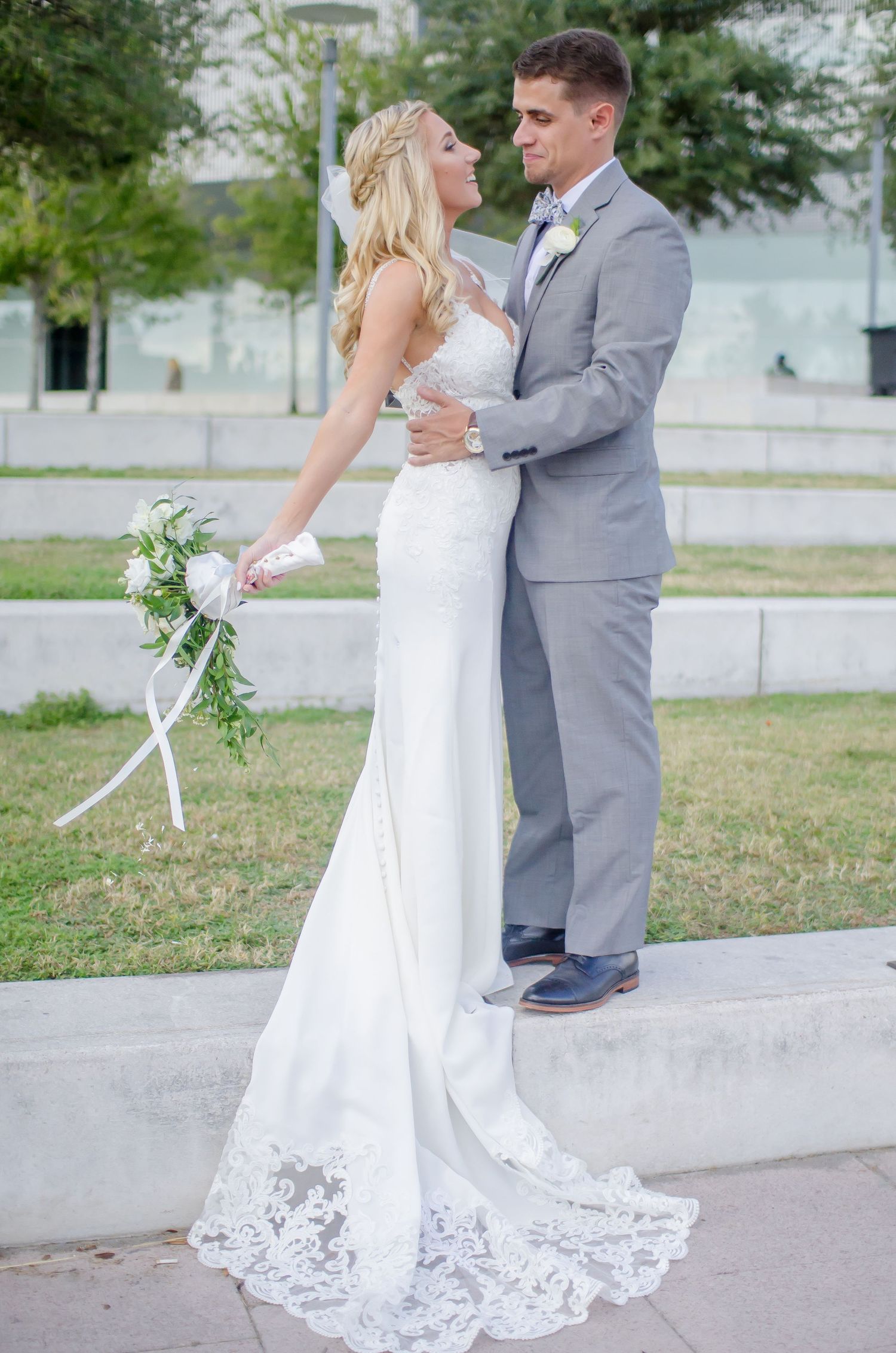 Bride in white lace gown holding bouquet embraces groom in gray suit outdoors on their wedding day.