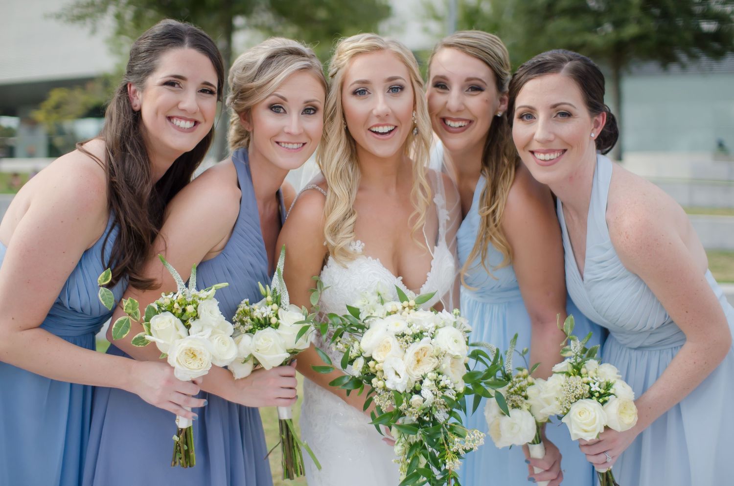 Bride with four bridesmaids in blue dresses holding white floral bouquets pose together outdoors.