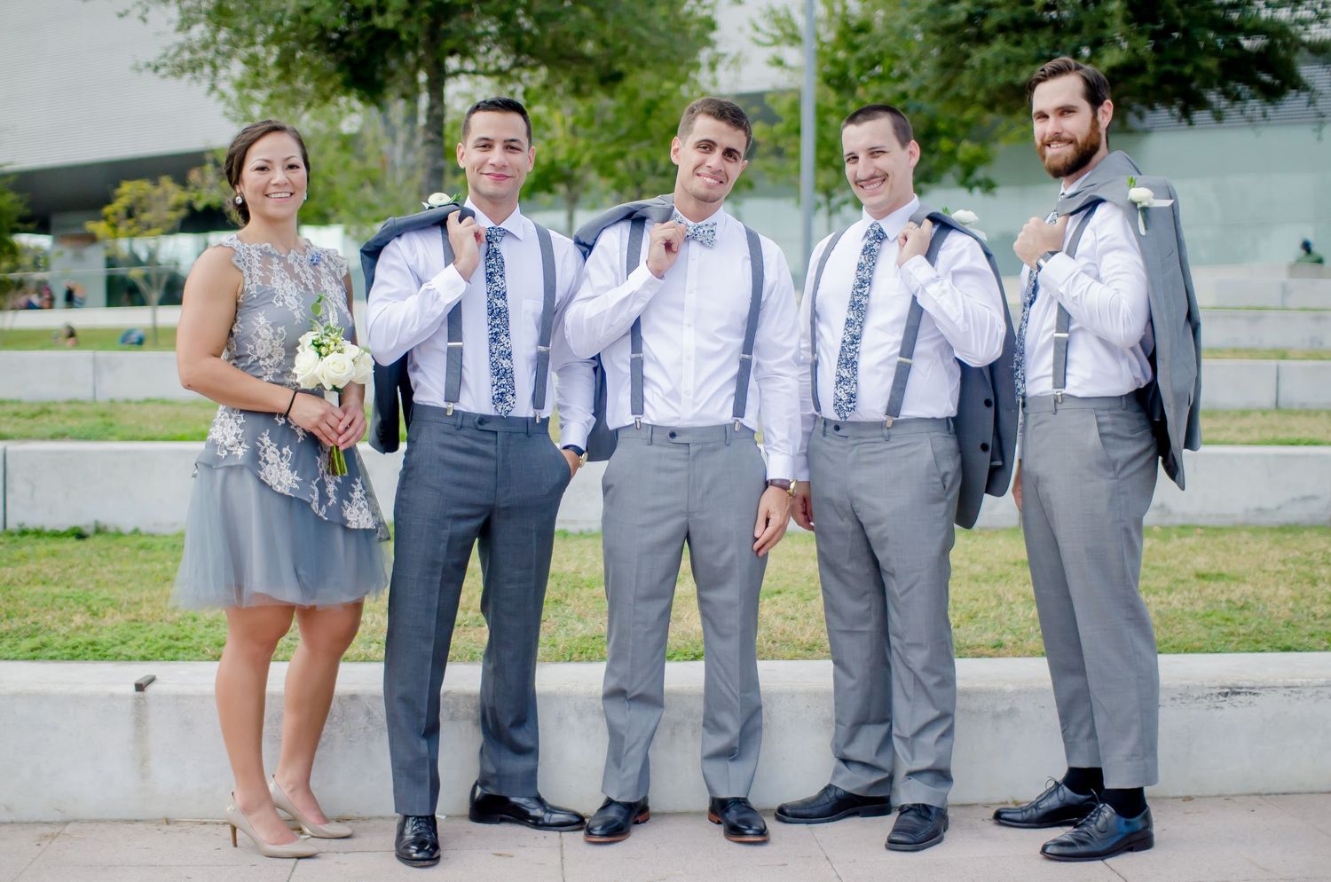 Groomsmen in gray suits with suspenders pose with bridesmaid holding bouquet outdoors at wedding.