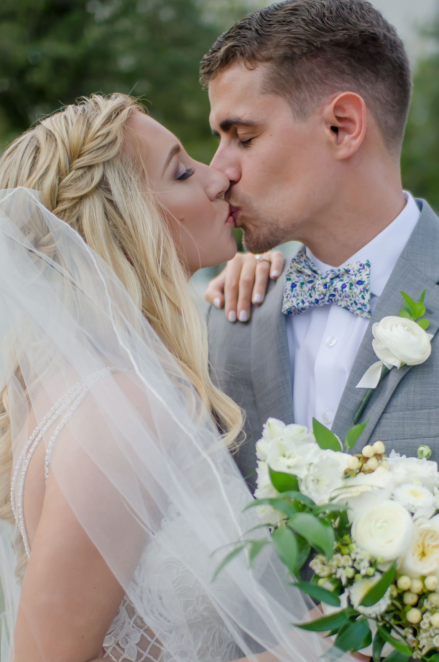 Bride and groom sharing a romantic kiss, bride holding white floral bouquet, groom wearing gray suit with floral bow tie.