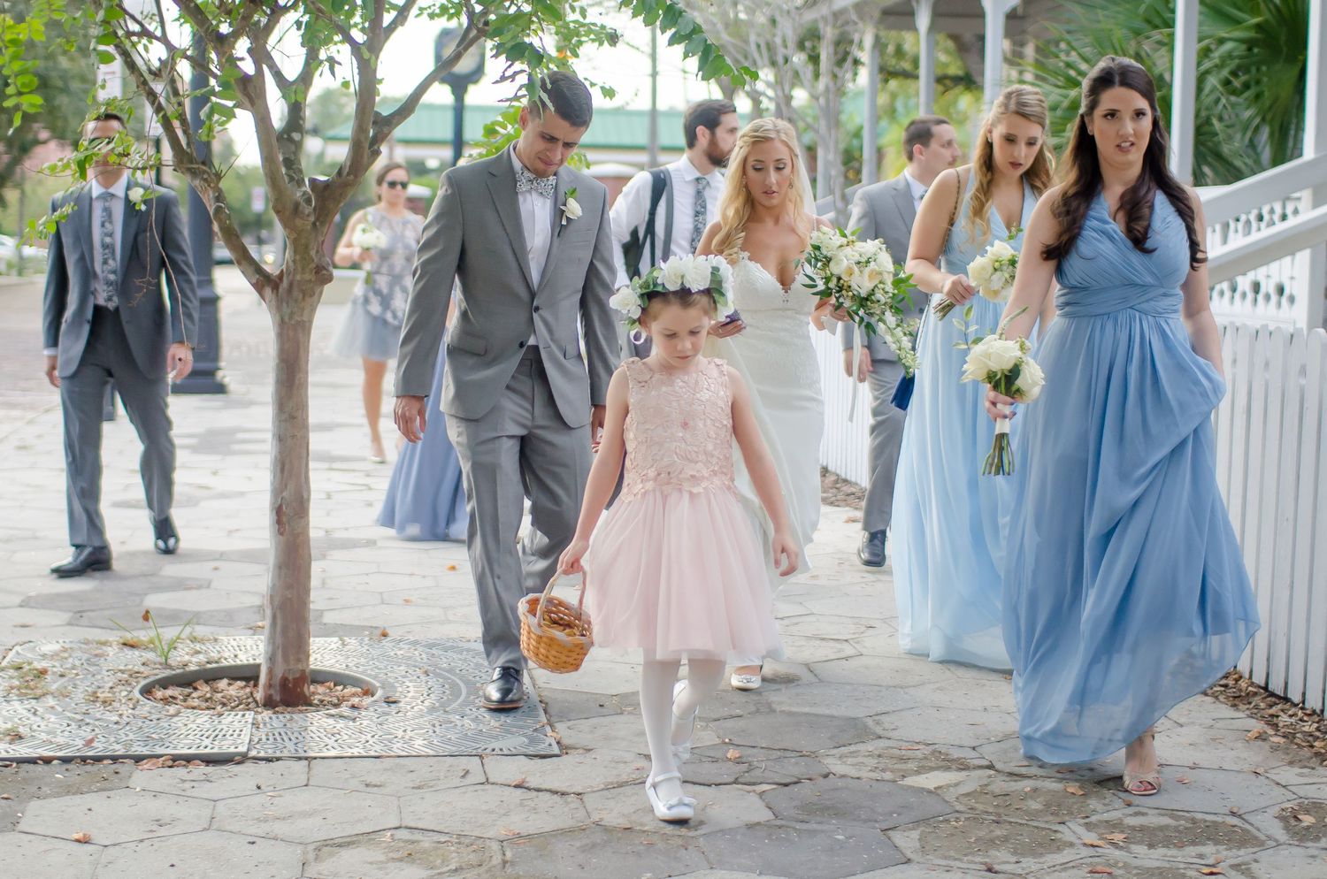 A flower girl in pink leads a wedding procession with bridesmaids in blue and groomsmen in gray suits outdoors.
