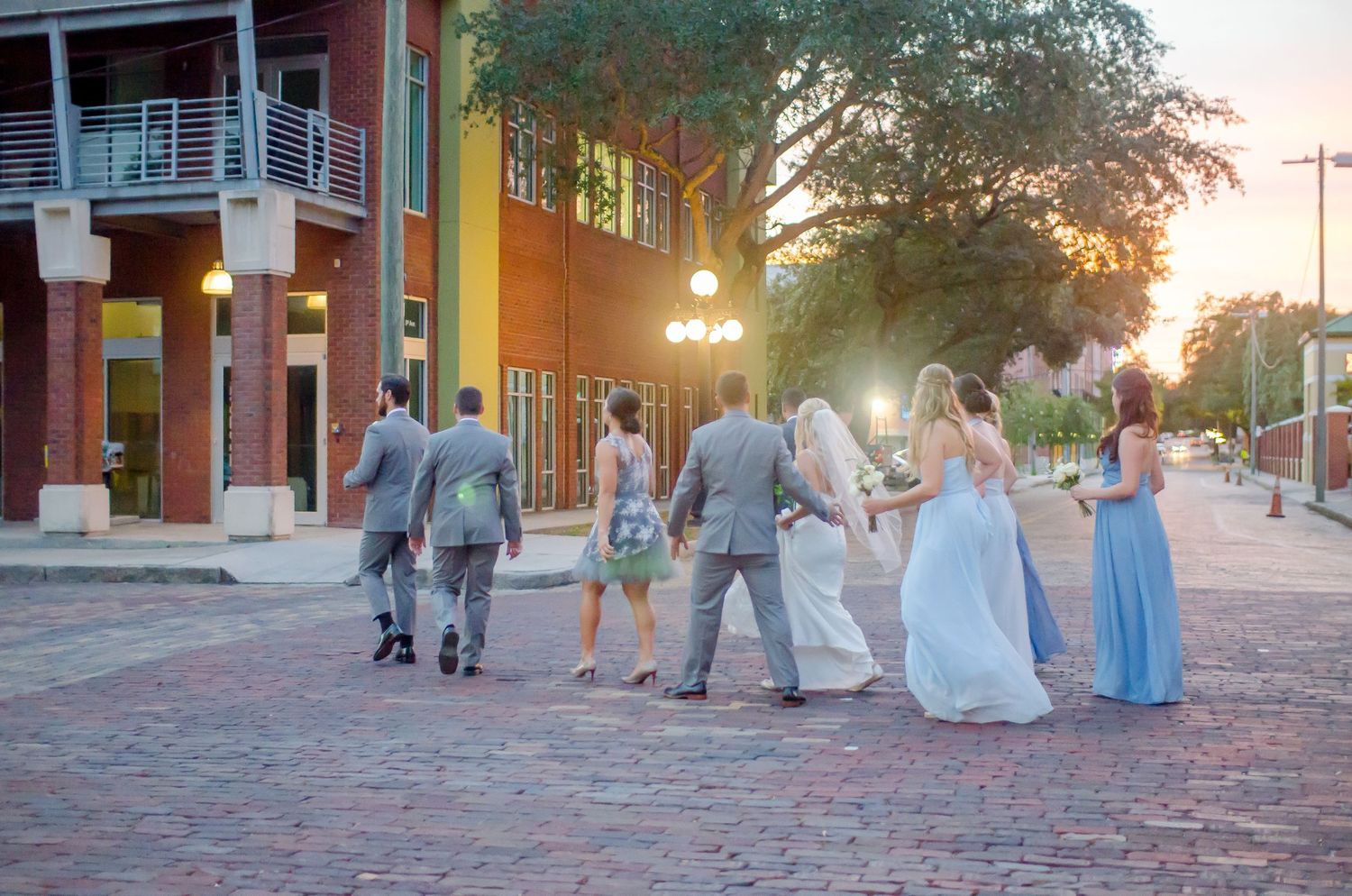 Wedding party walks on cobblestone street at sunset near brick buildings, bridesmaids in blue gowns.
