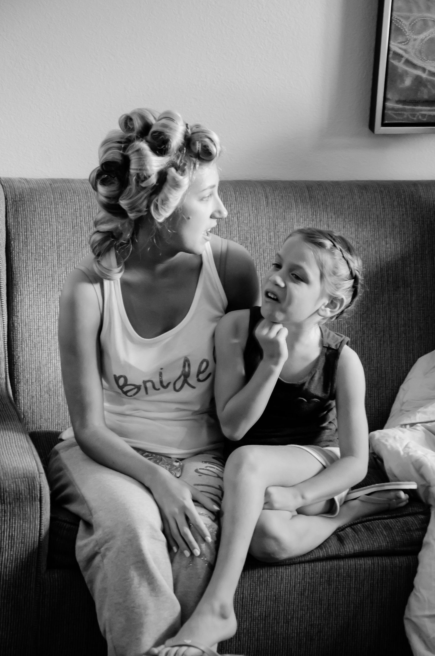 Black and white photo of a bride with hair rollers in a tank top sitting on a couch with a young girl.