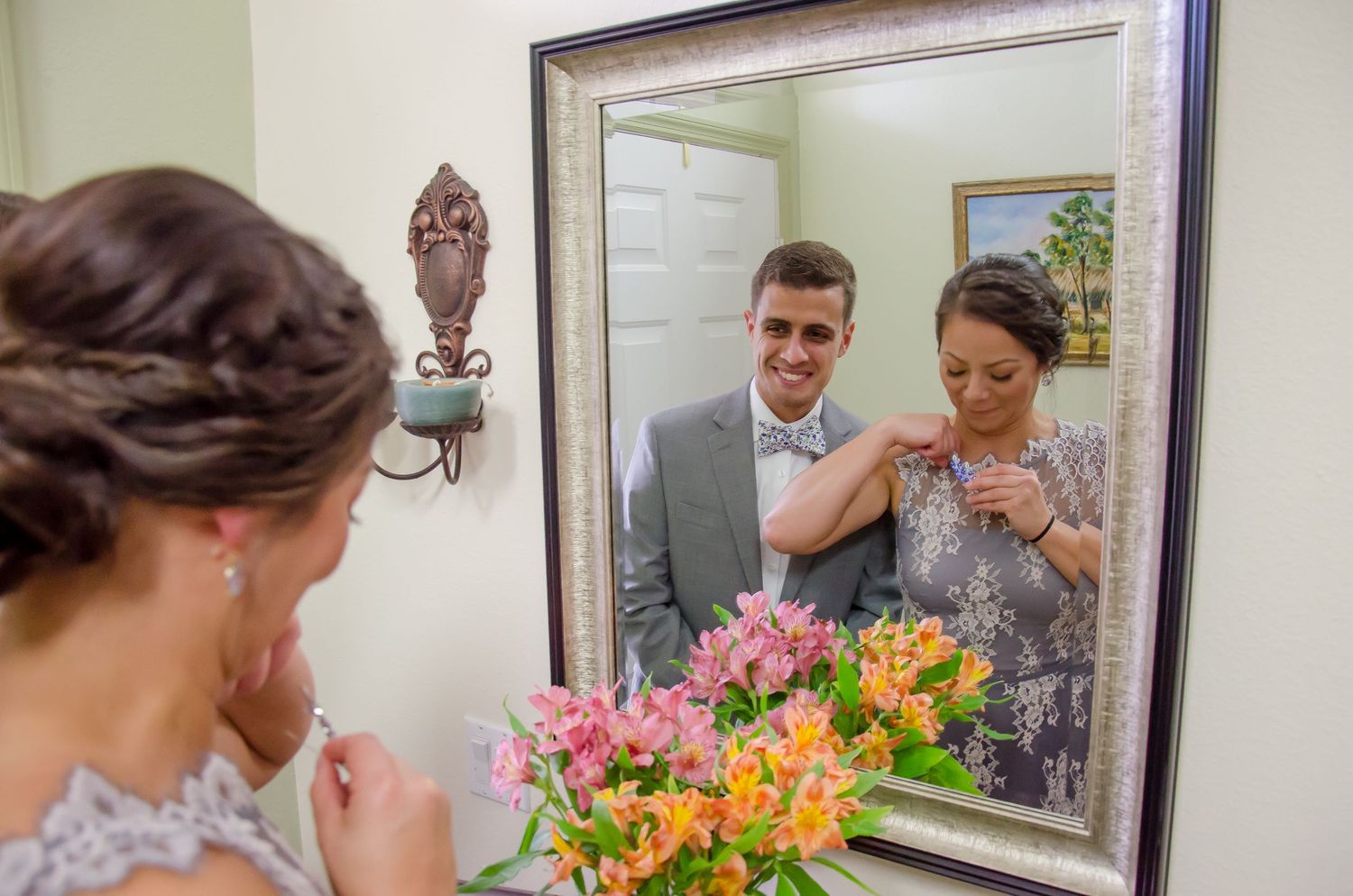 Bride adjusting groom's bow tie reflected in ornate mirror with colorful flowers in foreground at wedding venue.