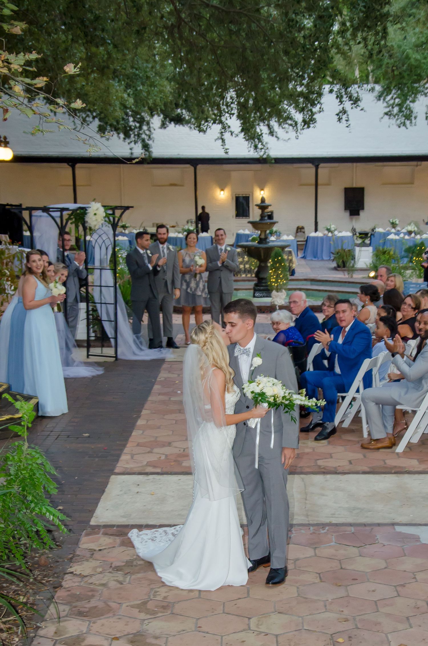 Bride and groom share first kiss as newlyweds at outdoor wedding ceremony with guests applauding.