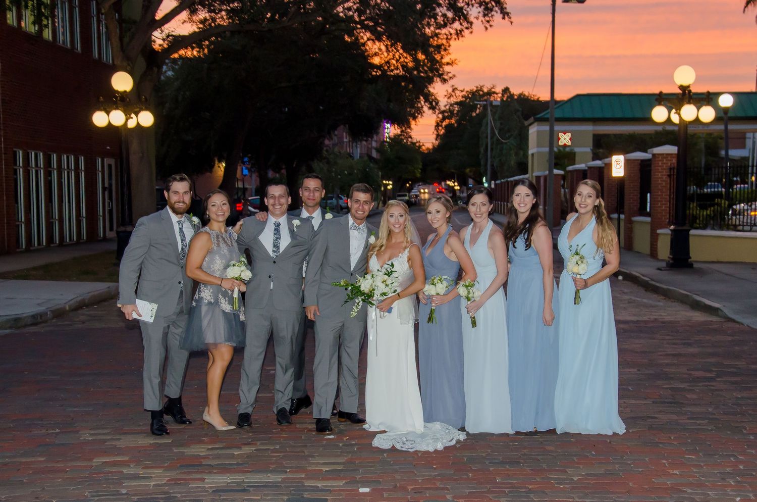 Wedding party poses at sunset on brick street, bride in white gown with groomsmen in gray suits and bridesmaids in blue dresses.