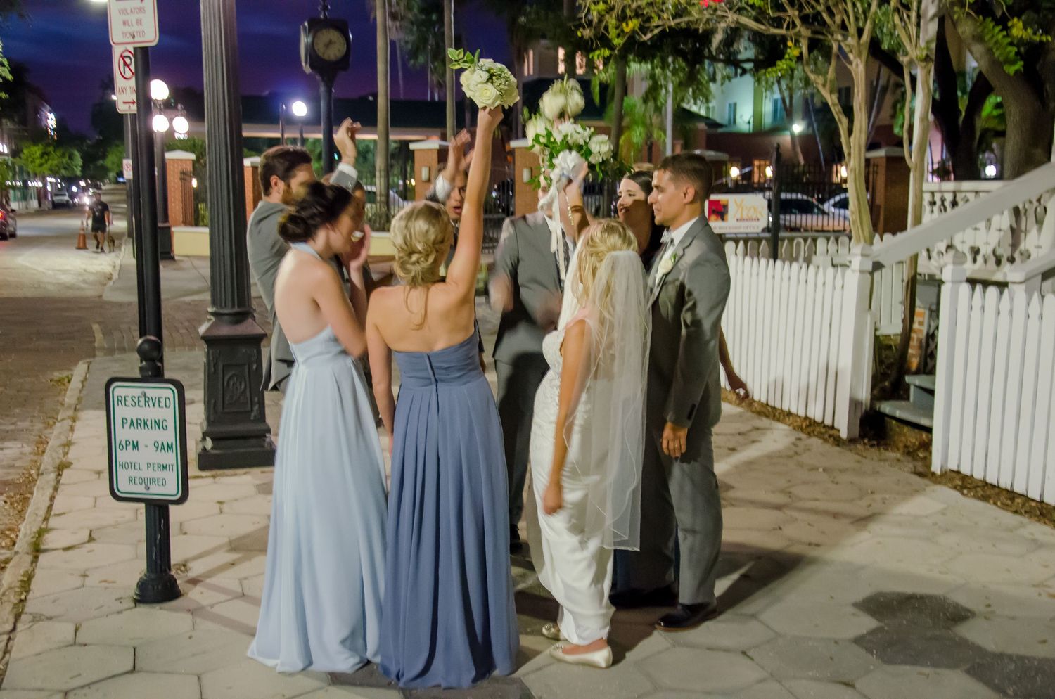 Wedding party in blue gowns and suits gathered on a sidewalk at night, bride holding bouquet aloft.