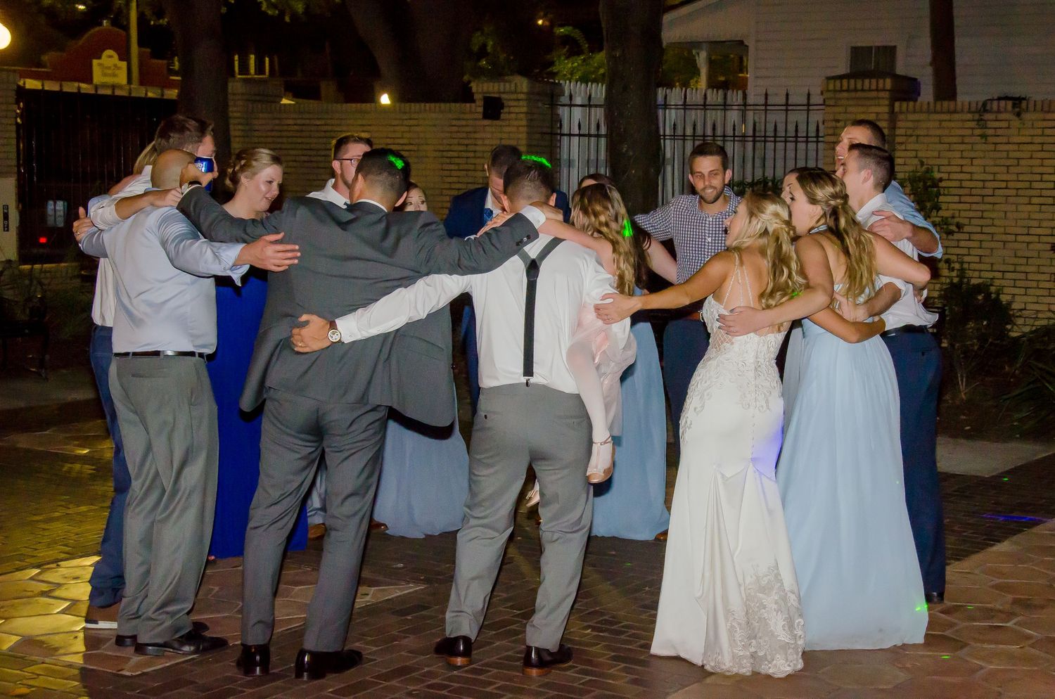 Wedding party dancing together outdoors at night, bride in white gown surrounded by guests in formal attire.