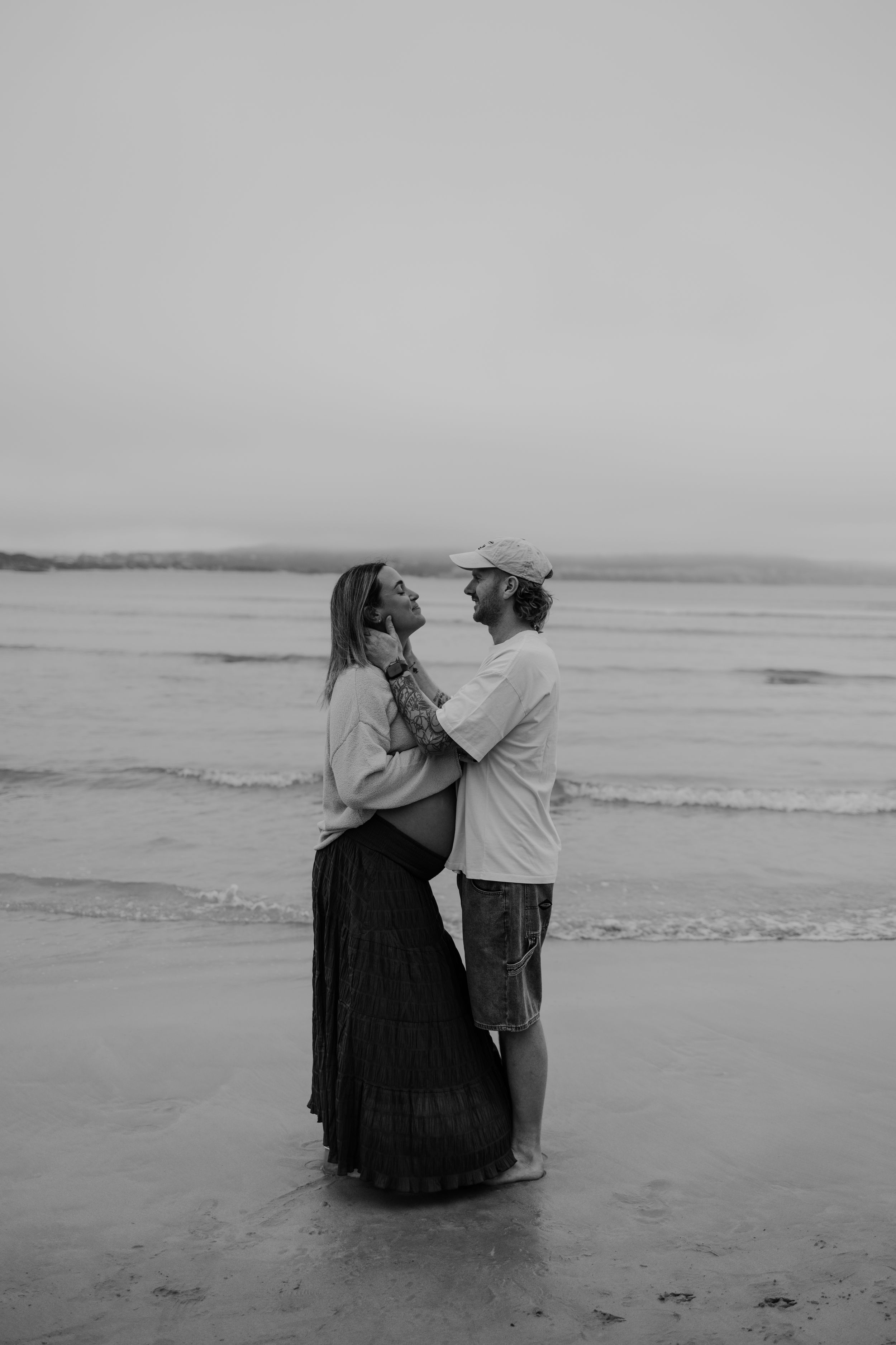 Black and white beach maternity photo of couple embracing by ocean waves, woman in flowing skirt showing baby bump.