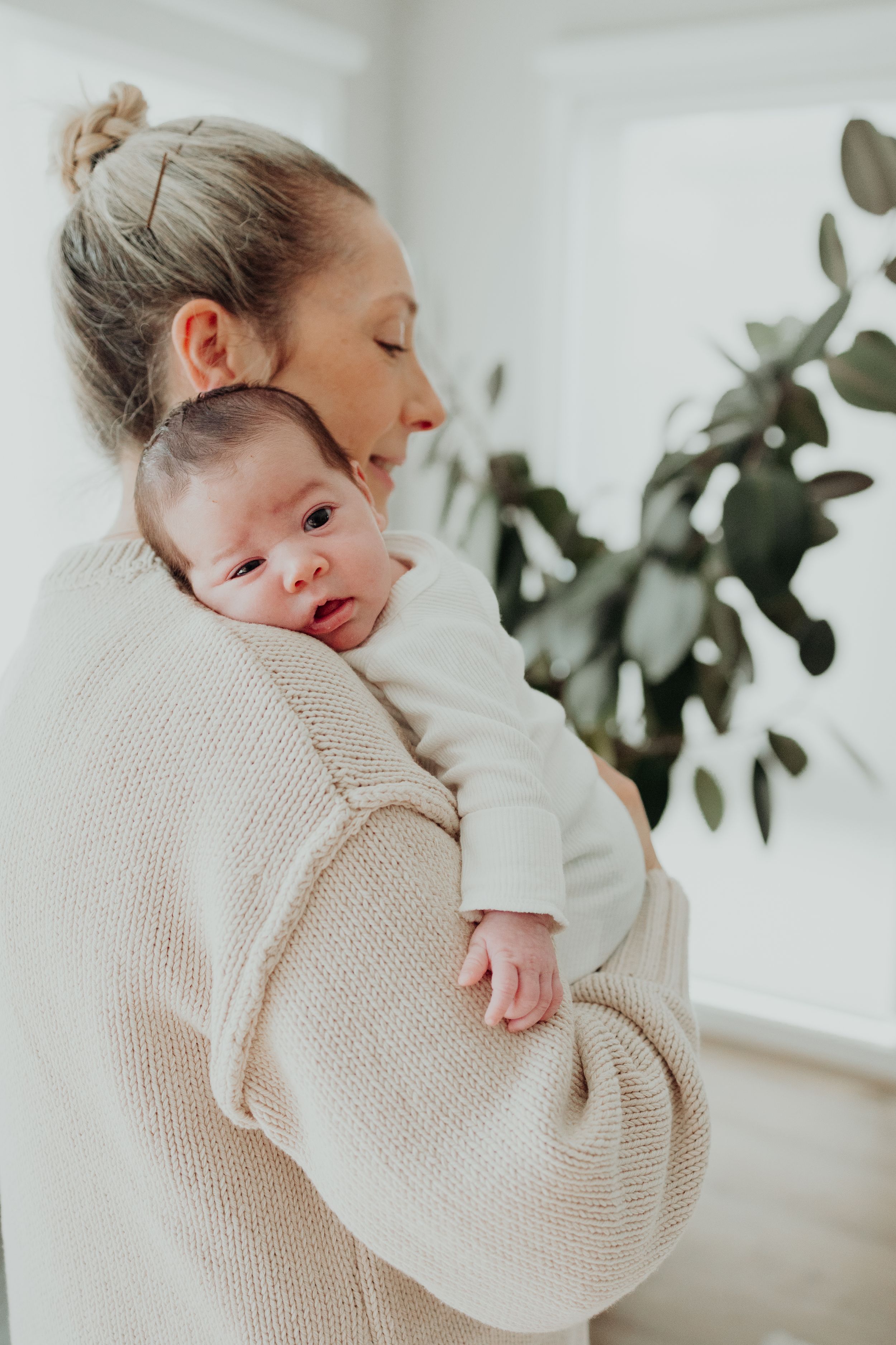 Blonde mother in cream knit sweater gently holding newborn baby against her shoulder indoors.