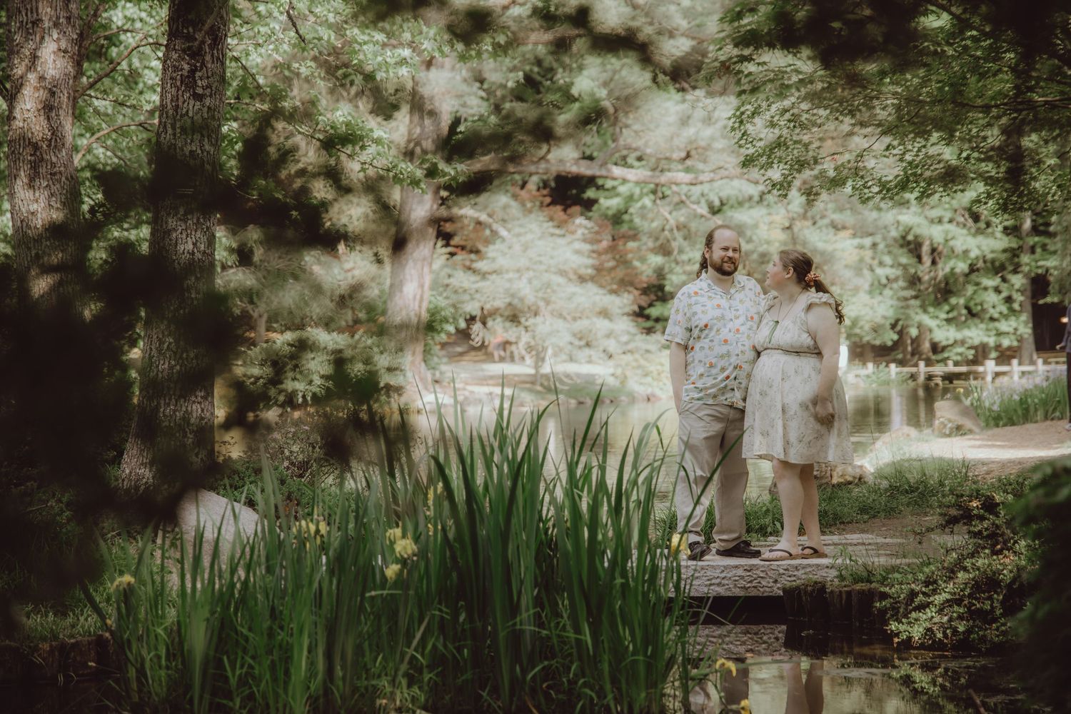 Couple holding hands on a wooden bridge surrounded by lush green trees and iris flowers in a serene garden setting.