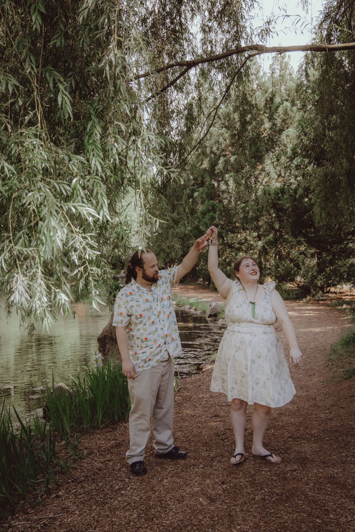 A couple joyfully holding hands under willow trees beside a tranquil pond in a lush green park setting.