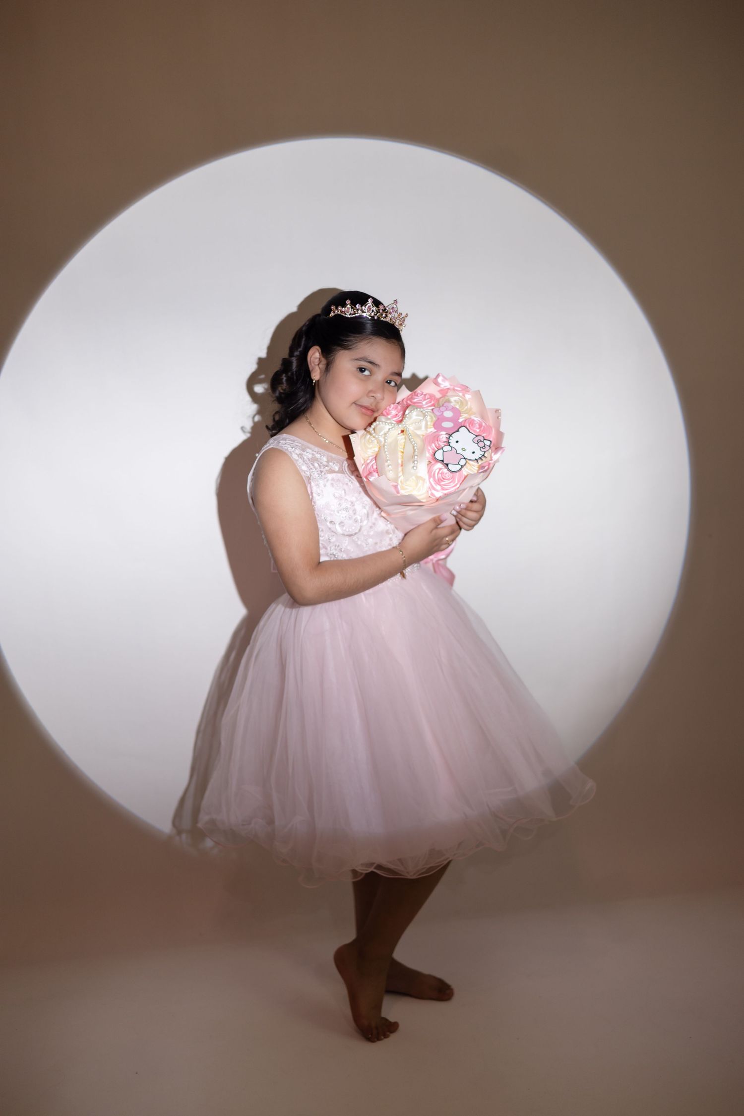 Young girl in pink tulle dress and tiara holds floral bouquet, posing in front of circular light backdrop.
