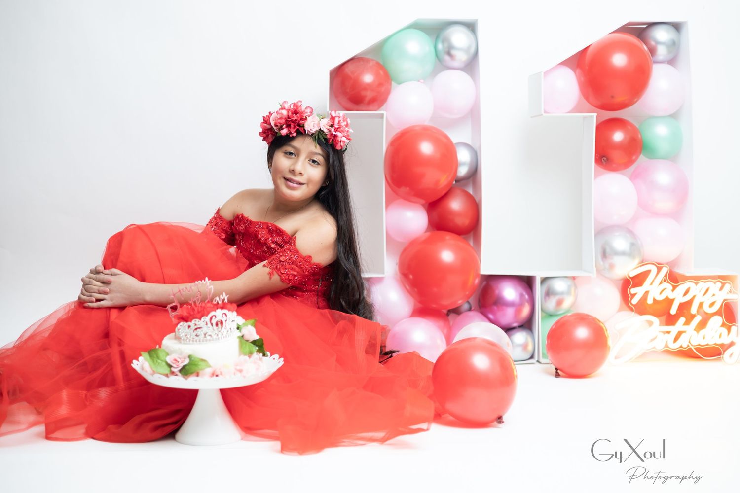 Girl in red gown celebrates 11th birthday with cake, balloons, and floral crown in a festive photo shoot.