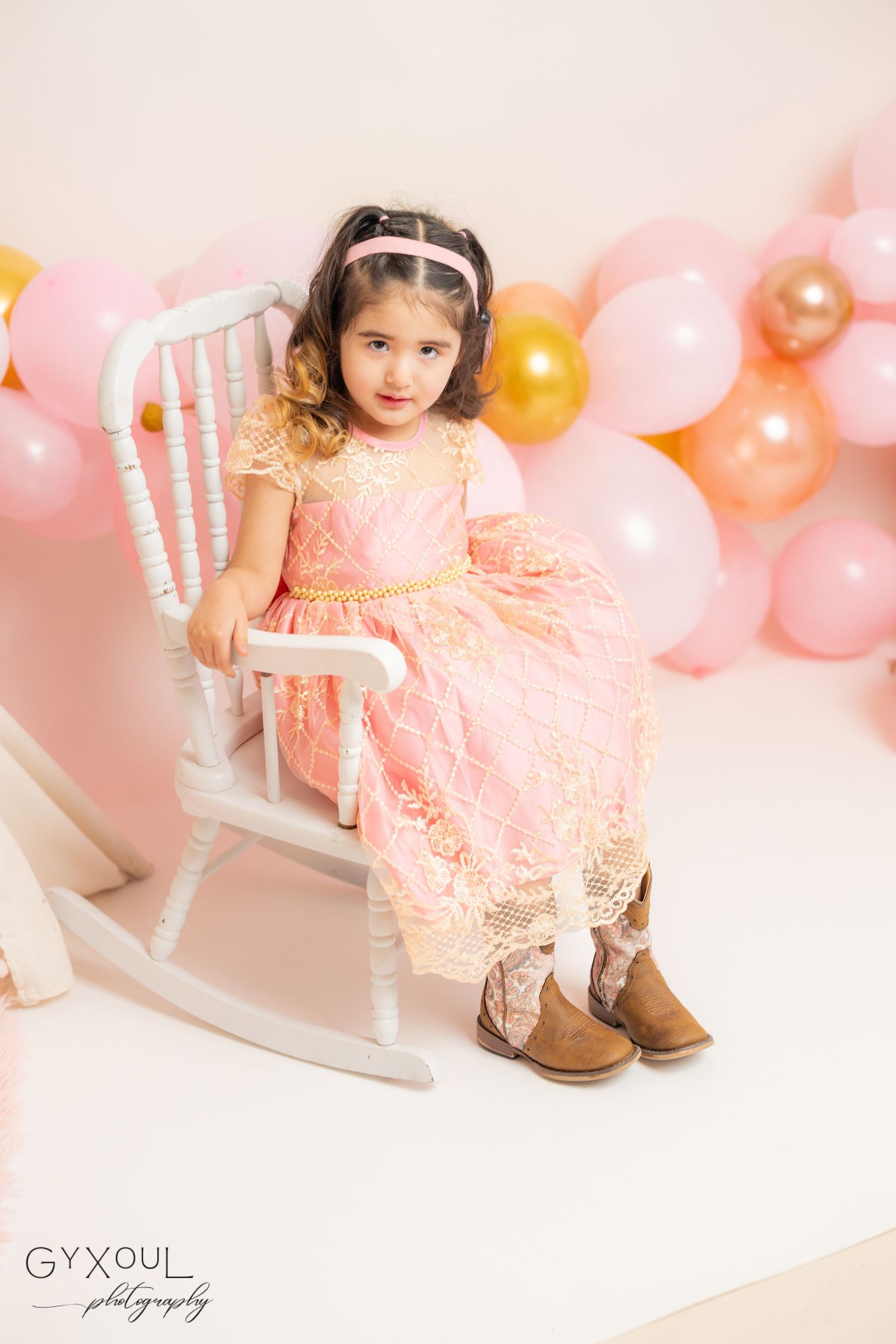 Young girl in pink lace dress and cowboy boots sits on white rocking chair with pink and gold balloon backdrop.
