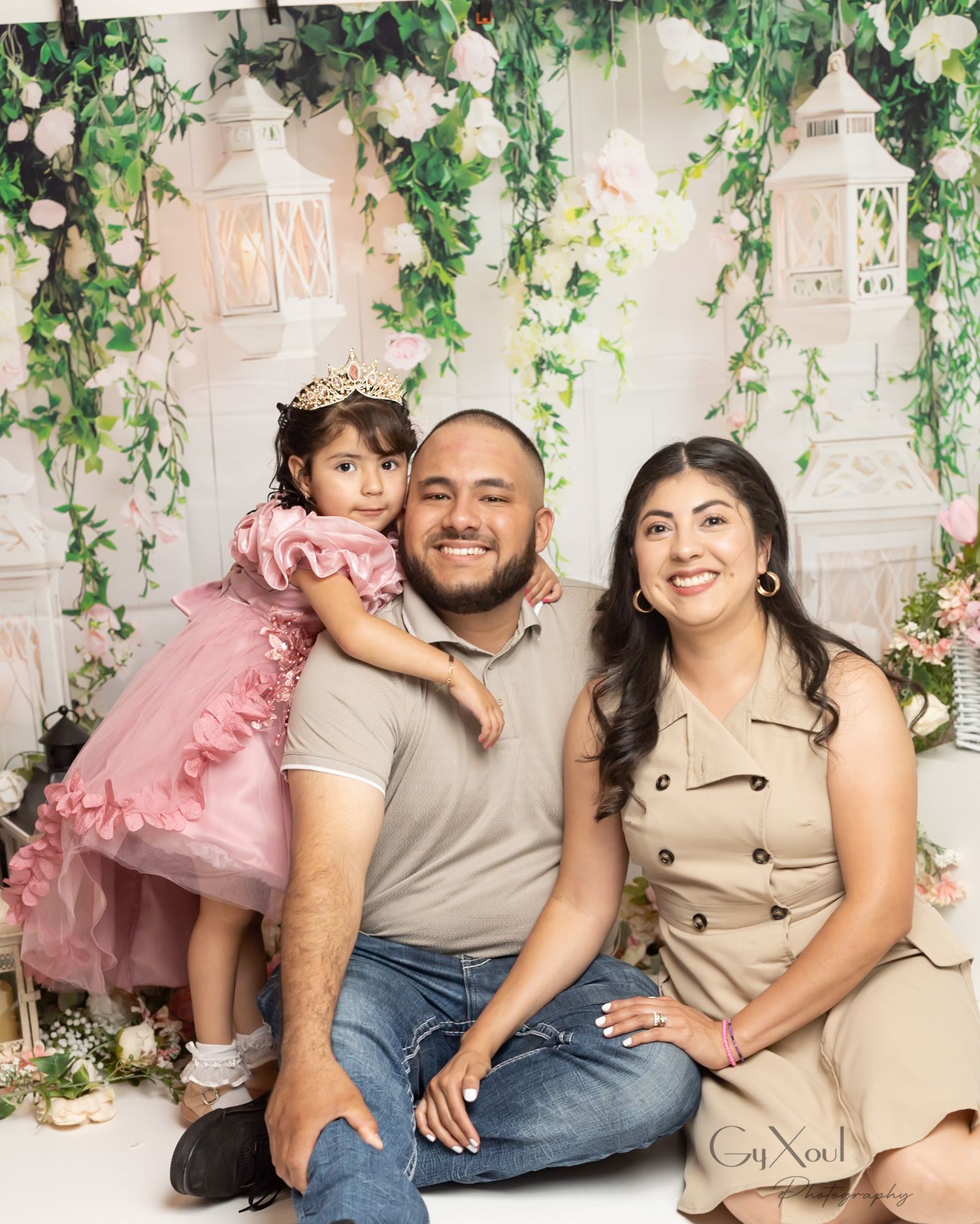 Family portrait with parents and young daughter in pink dress posing in front of floral backdrop with white lanterns.