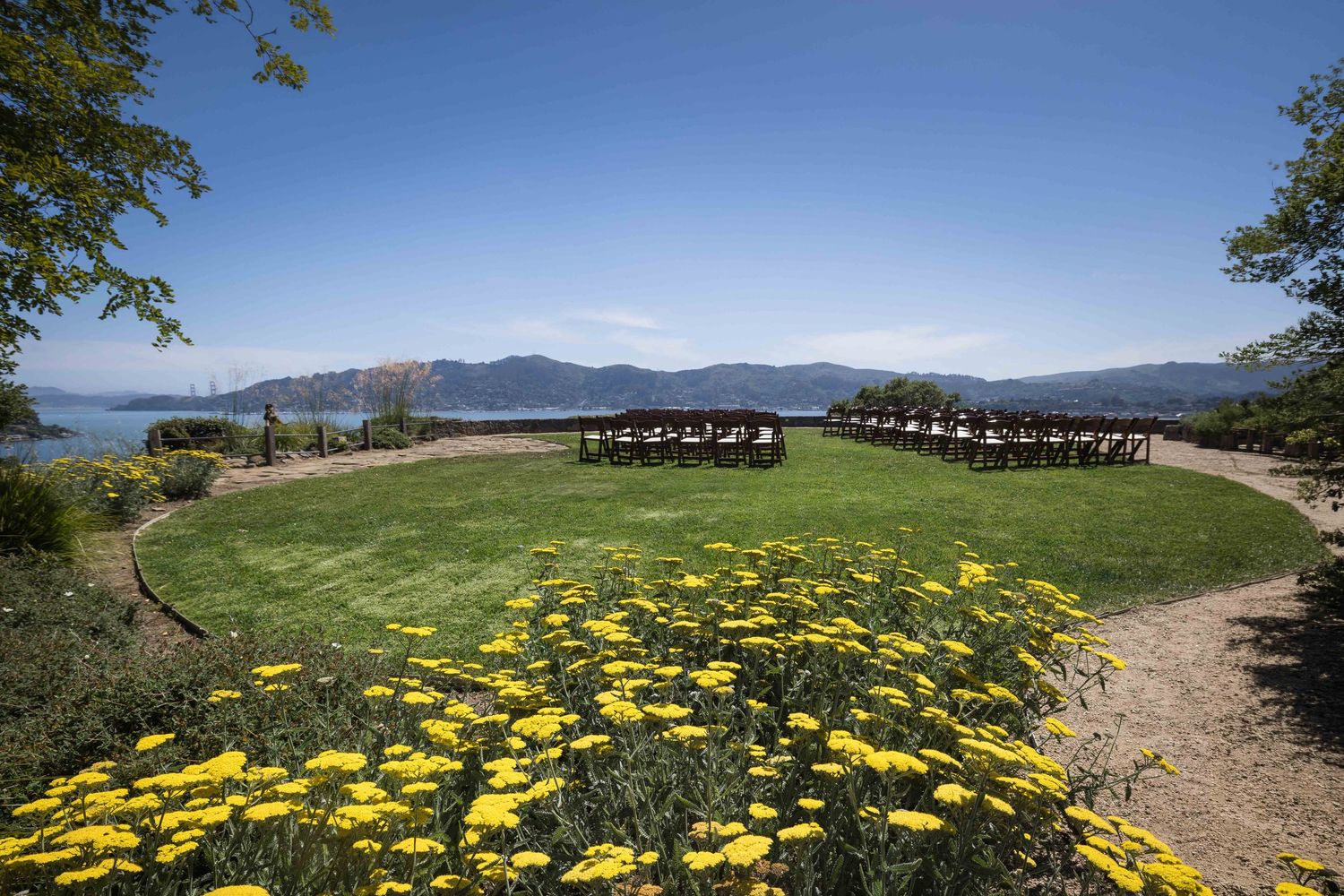 Outdoor wedding venue with yellow wildflowers, green lawn, mountain lake views, and arranged seating under clear blue sky.