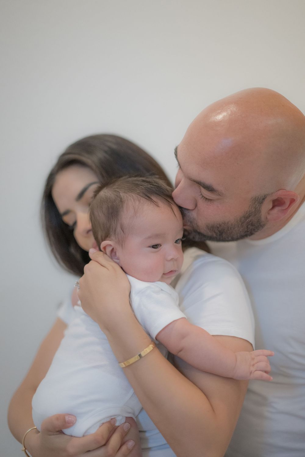 Bald father kissing newborn baby held by dark-haired mother, both wearing white shirts.
