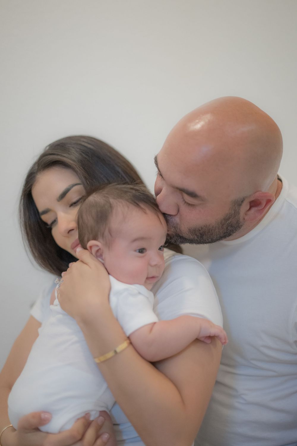 Loving parents in white shirts kiss and cuddle their newborn baby against a white background.