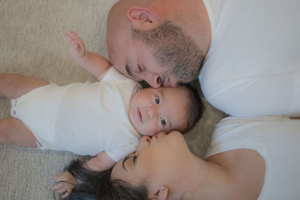 Two parents lying down kissing their smiling newborn baby on a soft carpet in a tender family portrait.