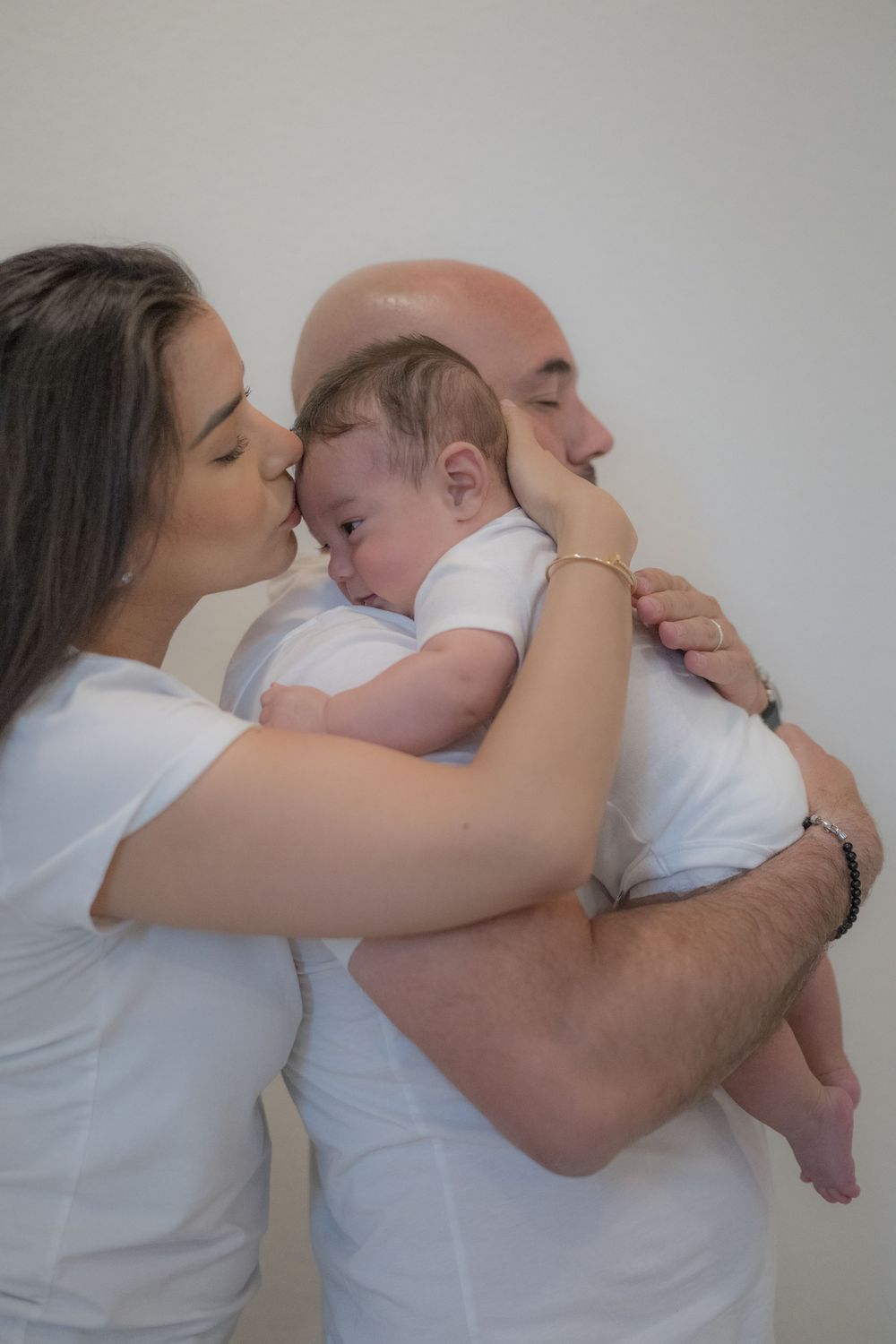 Dark-haired mother kissing bald father on cheek while he holds their sleeping newborn baby.