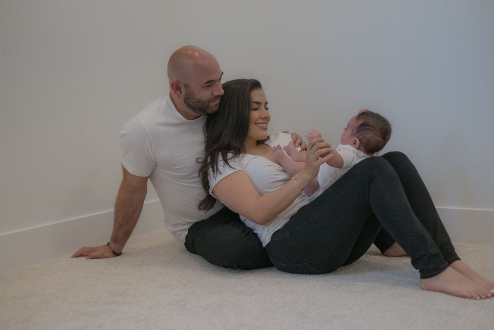 A couple sits on the floor smiling at their newborn baby in a bright, minimalist room.