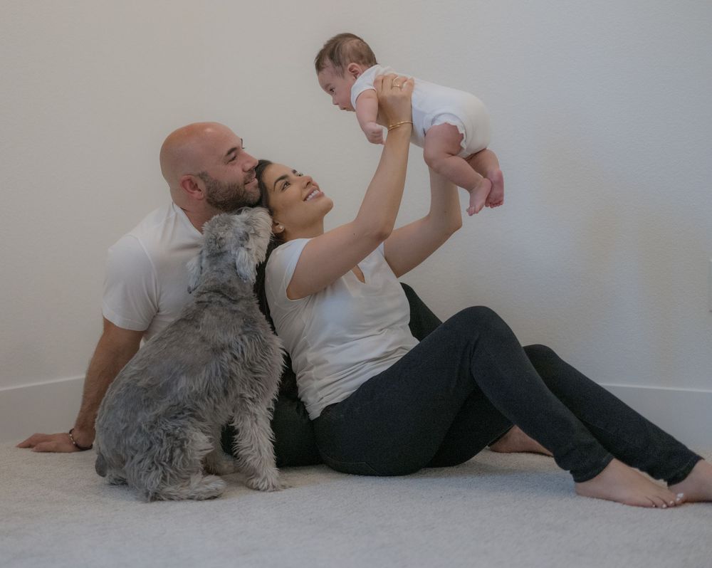 Family portrait with bald dad, mom lifting baby, and fluffy gray dog sitting together on floor.