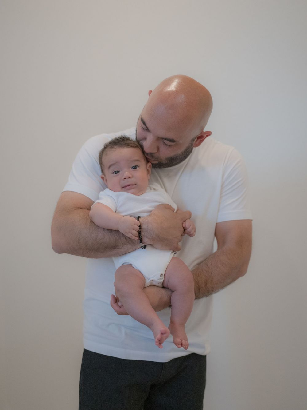 Bald man in white t-shirt tenderly kissing a baby dressed in white onesie while holding them close.