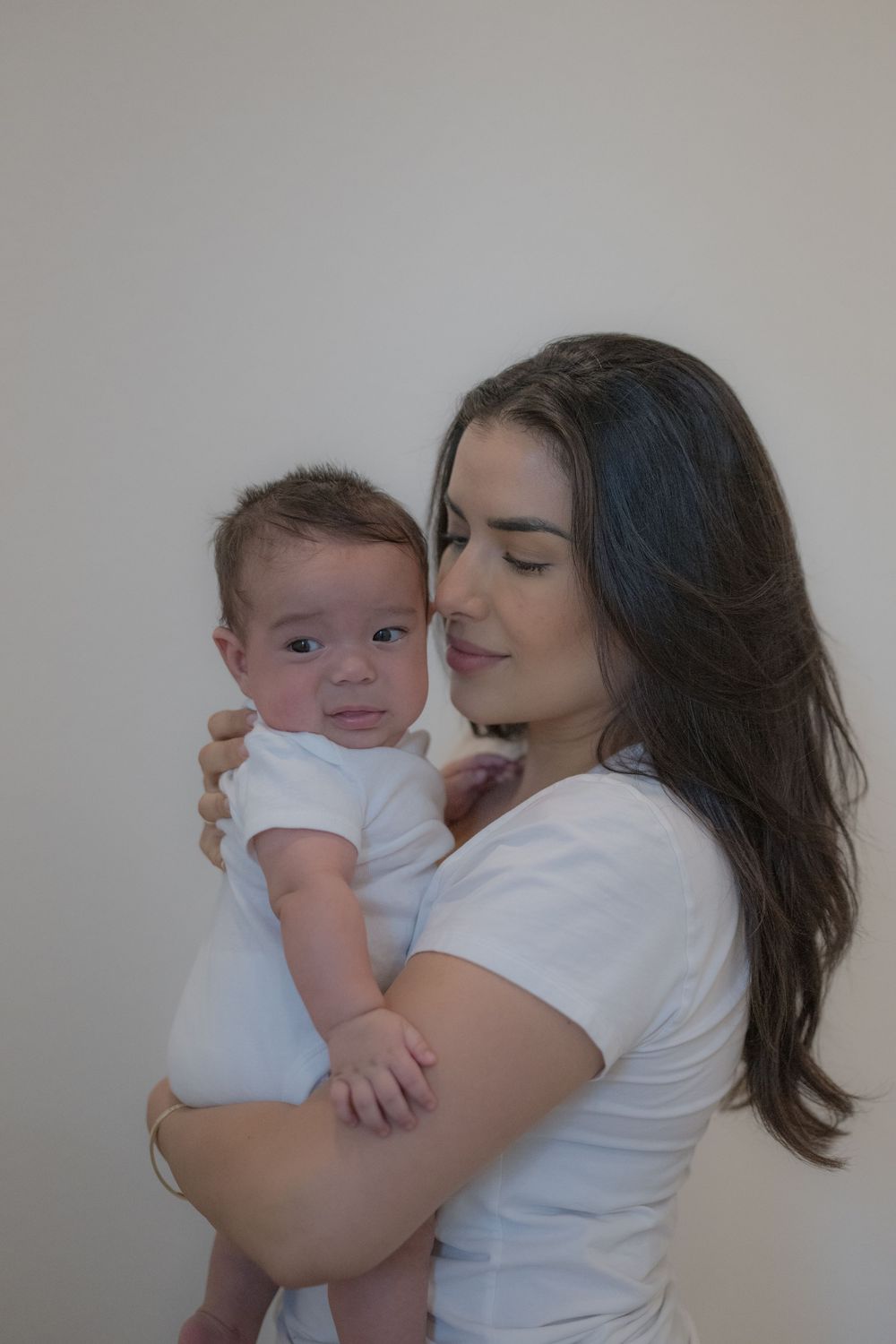 A young mother in a white shirt tenderly holds her newborn baby dressed in a white onesie.