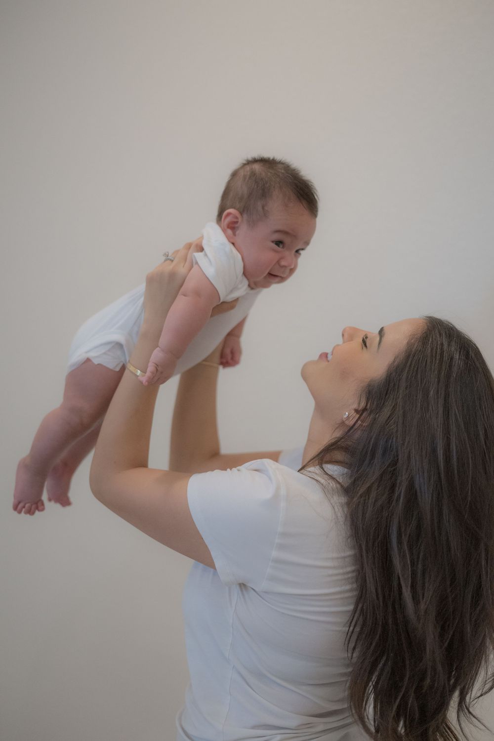 Mother in white shirt joyfully lifting smiling baby in white onesie above her head against neutral background.