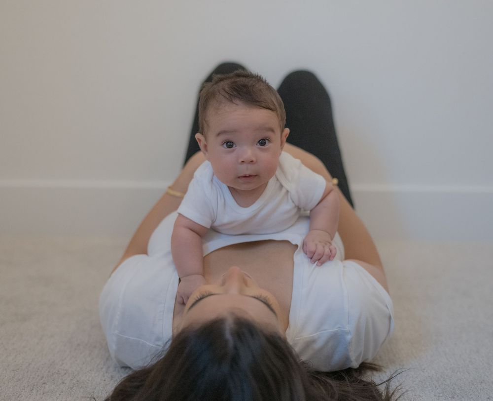 A mother lies on the floor as her newborn baby rests on her stomach, looking curiously at the camera.