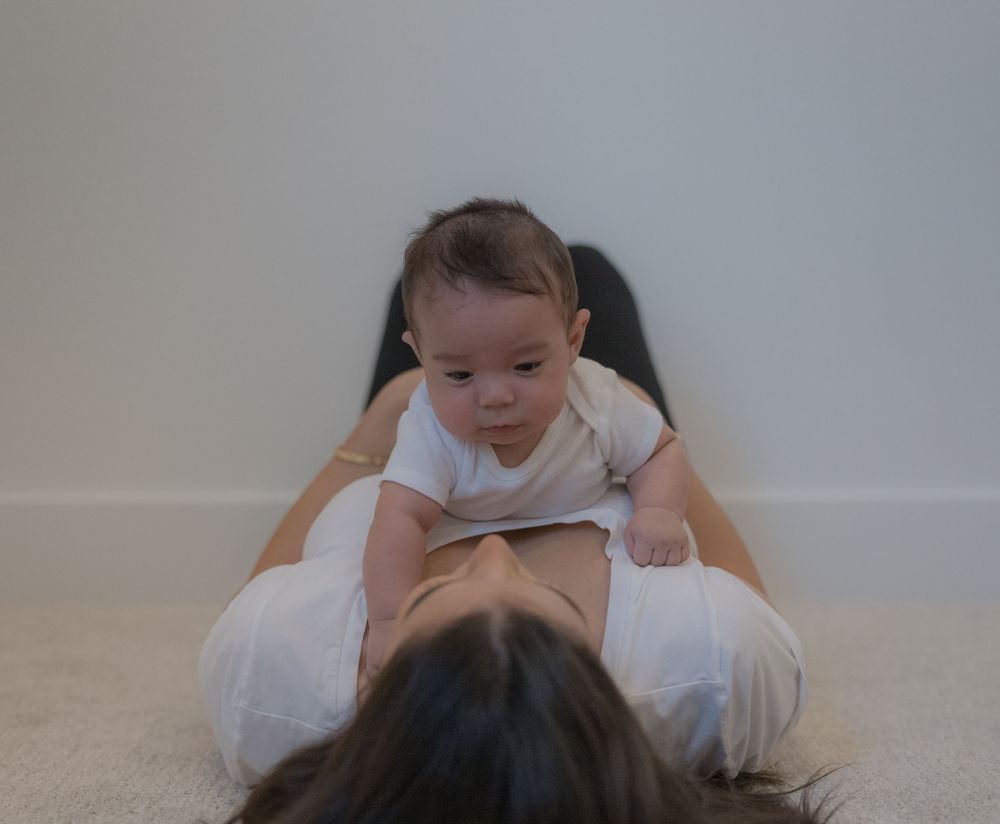 Mother lying on floor while curious baby in onesie practices tummy time on her chest.