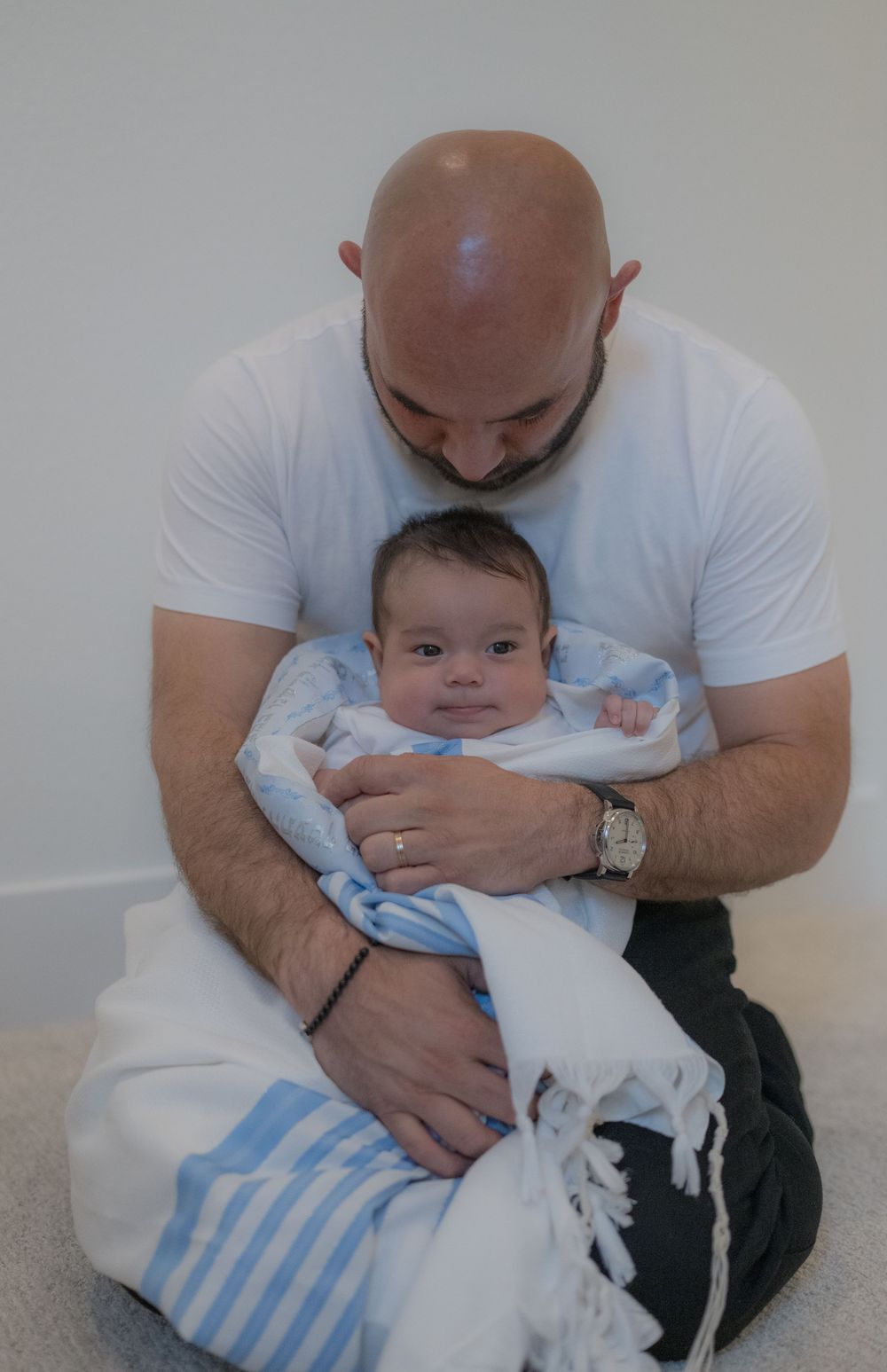 Bald father sitting cross-legged cradling newborn baby wrapped in blue and white striped blanket.