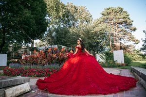 Young woman in a stunning red ball gown with long train poses in a lush garden for a quinceañera photo.