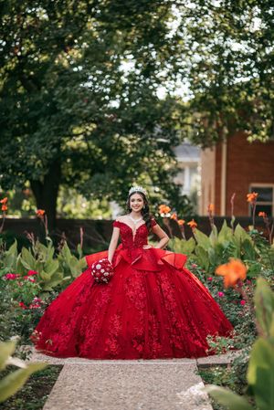 Young woman in red quinceañera ball gown with lace details and tiara poses in a garden holding a red bouquet.