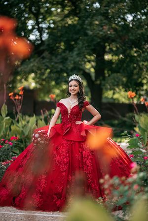 Smiling teen in red off-shoulder ball gown and tiara poses among colorful garden flowers for quinceañera photos.
