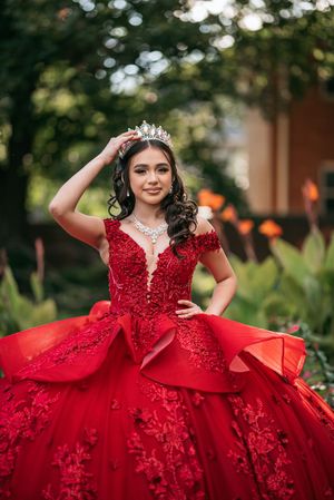 Young woman in red ball gown with lace detail and tiara poses outdoors for quinceañera portrait.