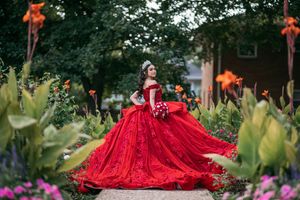 Young woman in a stunning red ball gown with lace details poses in a lush garden wearing a tiara.