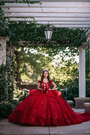 Young woman in a stunning red ball gown with lace details and tiara poses under a pergola with lush greenery.