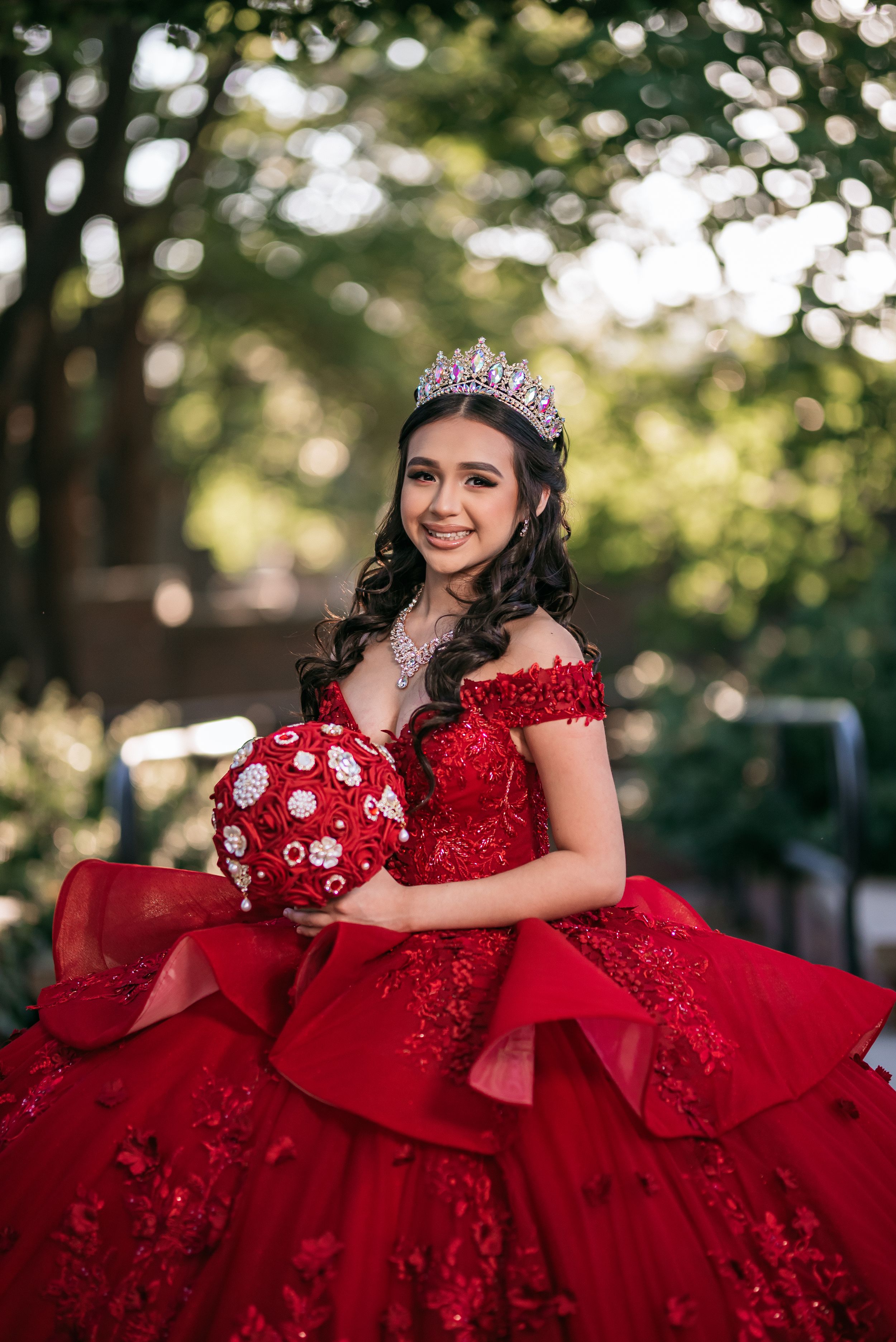 Young woman in red quinceañera ball gown with tiara holds floral bouquet, smiling outdoors among green trees.