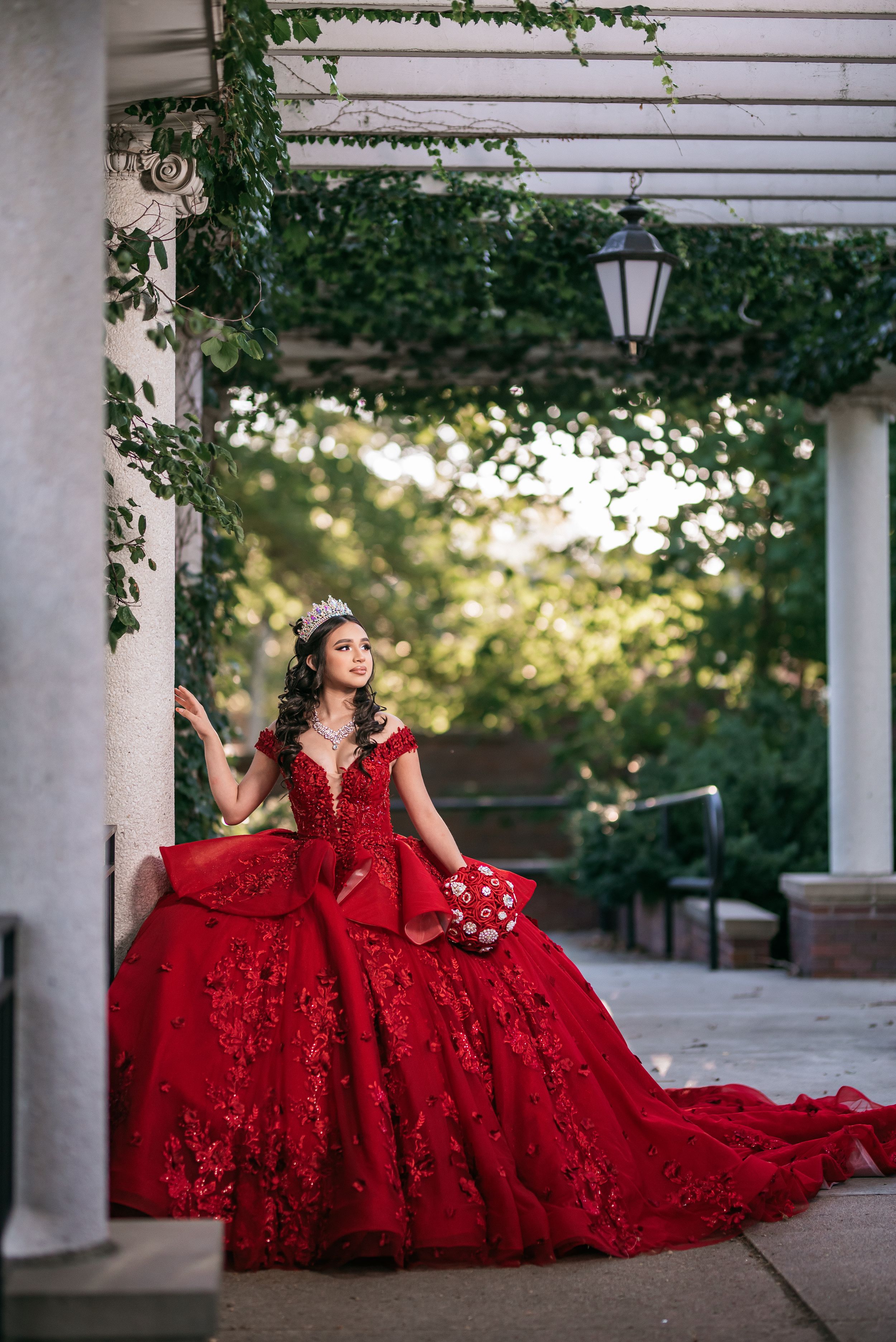 Young woman in red lace ball gown with crown poses under pergola surrounded by lush greenery for quinceañera.
