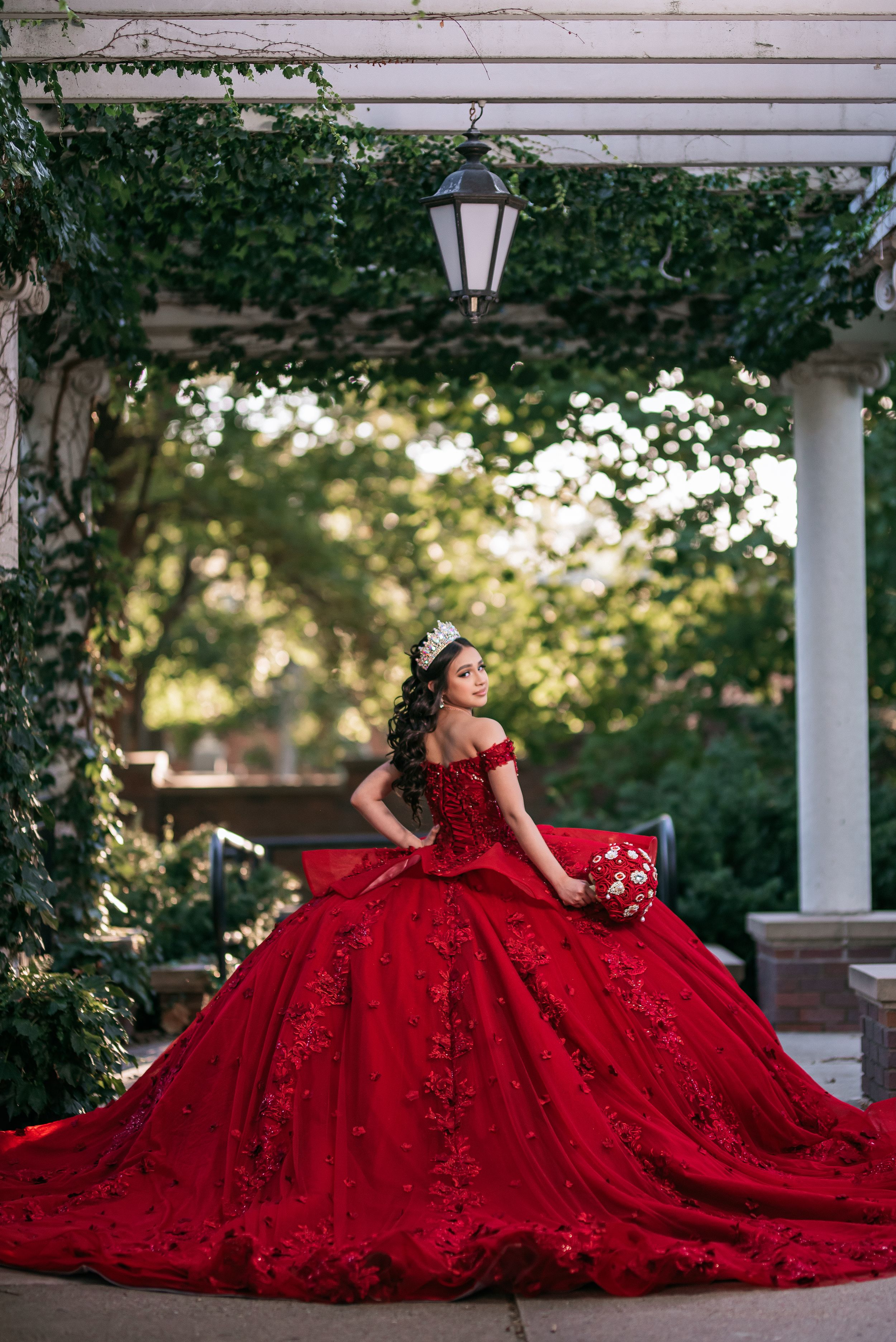 Young woman in a red ball gown with lace, wearing a tiara, holding a bouquet in an elegant garden setting.