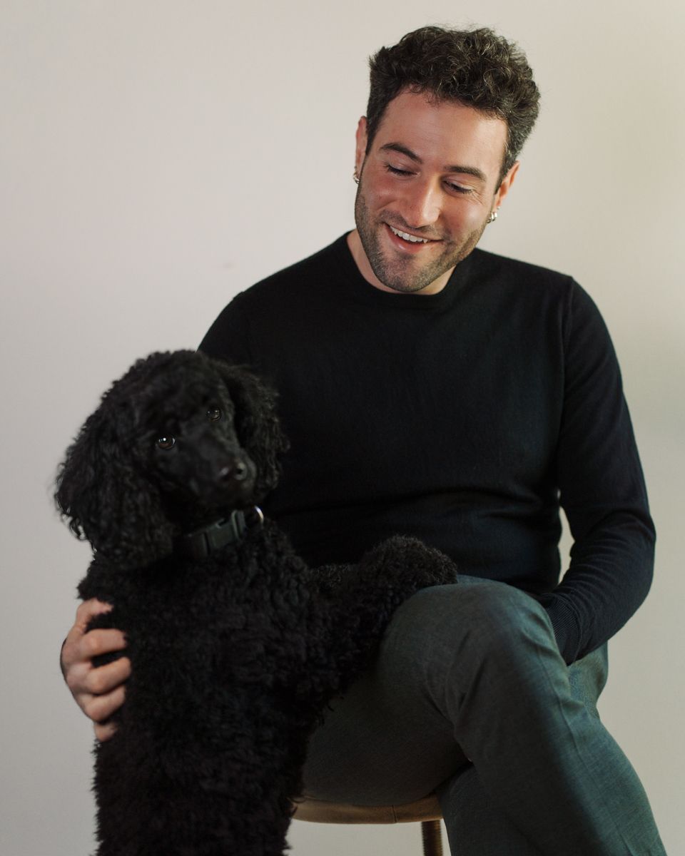 Smiling young man in black shirt holding a black poodle puppy while seated against a light background Sydney dog photoshoot in Hills distric