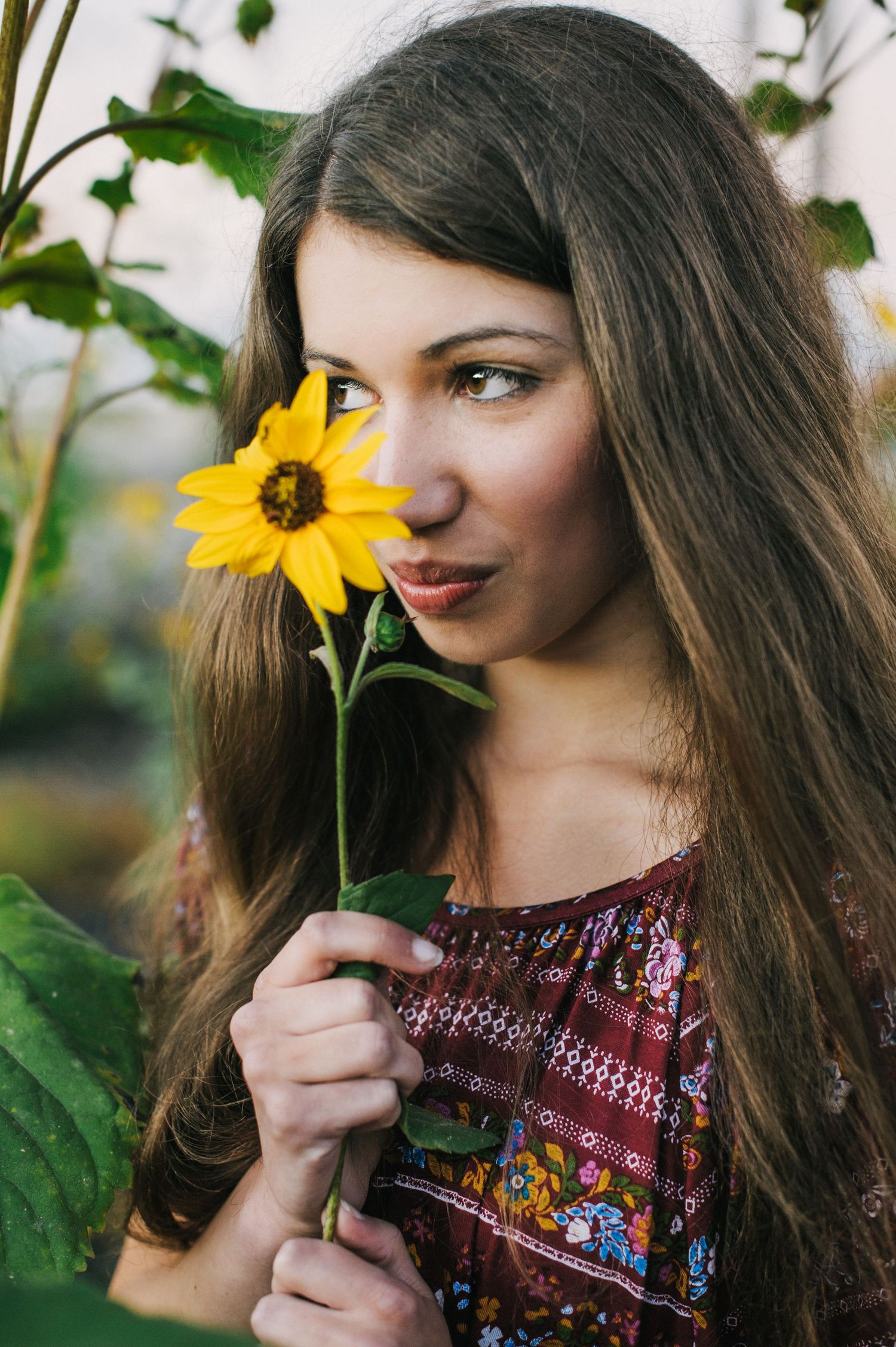Young woman with long brown hair holding a yellow sunflower near her face, wearing a floral dress outdoors.