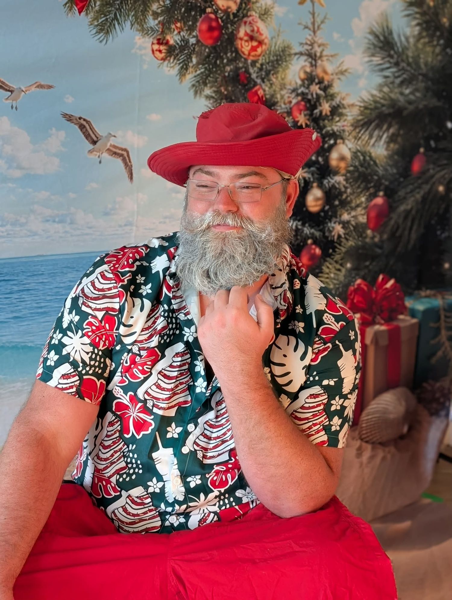 Man with gray beard wearing red cowboy hat and tropical Christmas shirt poses in front of holiday beach backdrop.