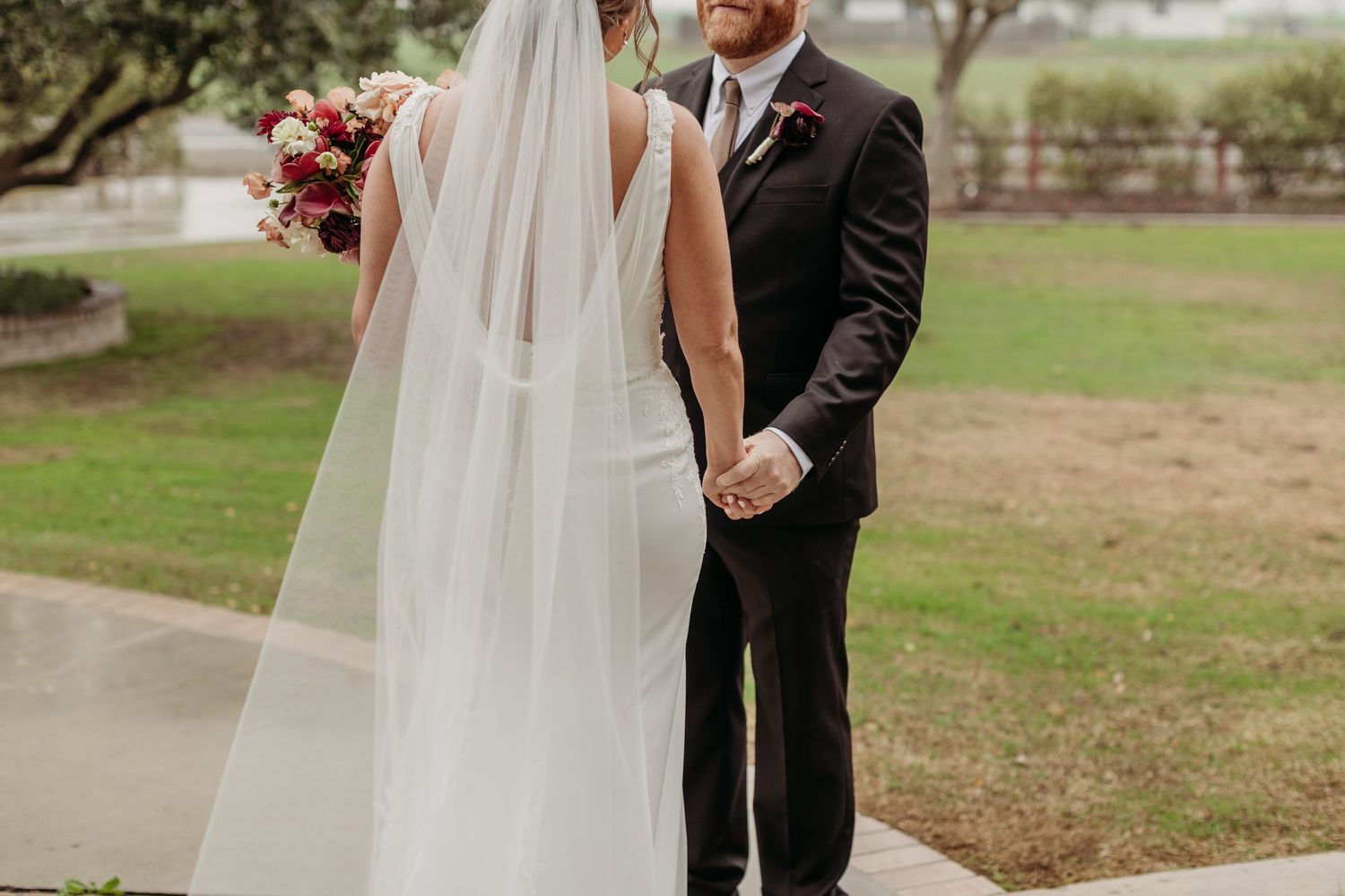 Bride and groom walking through lush green field, bride in elegant white gown holding bouquet, cloudy sky backdrop.